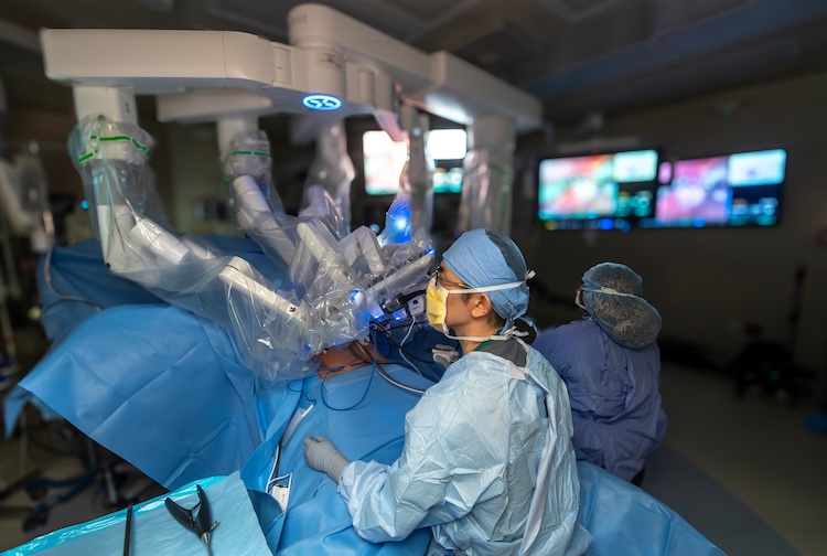 Surgeon sits at the side of an operating table with robotic arms above him and the patient.