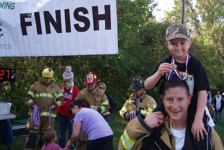 Greg Leitz holds up a small child after they completed a race. He is wearing his firefighter gear