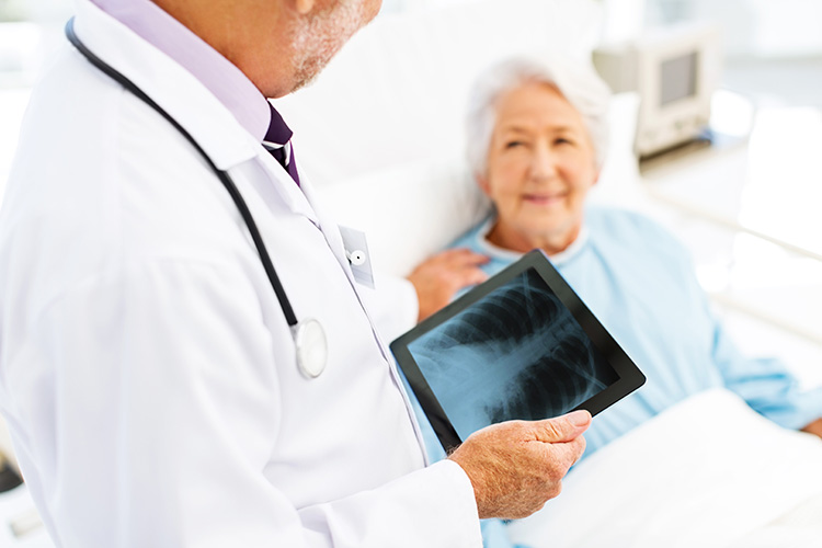 A physician viewing an x-ray of lungs and a patient in bed, in the background.