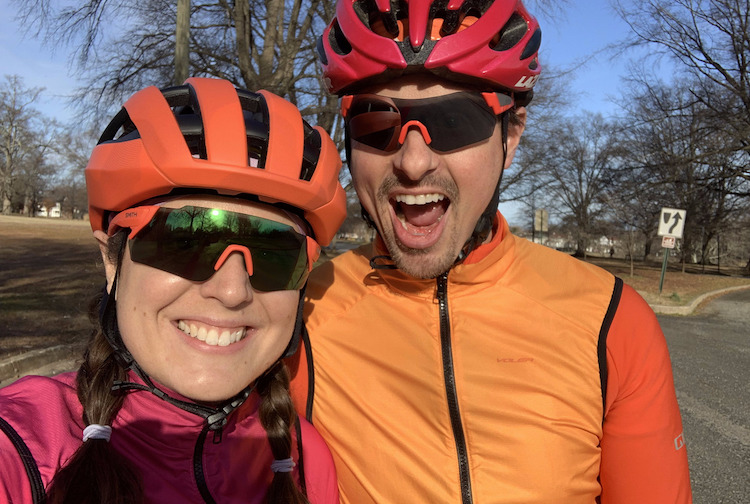 Woman and man smiling at the camera. They are wearing helmets and athletic gear for riding their bicycles.
