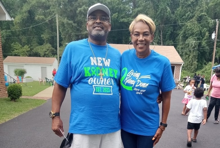 Two people standing in a neighborhood with blue shirts, one says that they received a transplant and the other says they're the donor